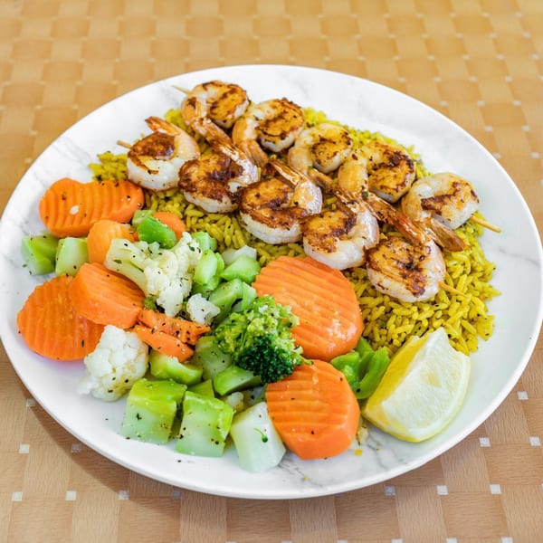 Grilled shrimp skewers on yellow rice with steamed carrots, broccoli, and cauliflower, garnished with a lemon wedge.